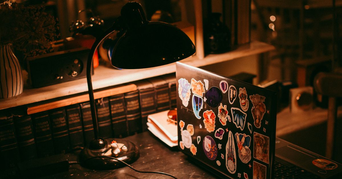 Warm lighting illuminates a workspace with a laptop, lamp, and books.