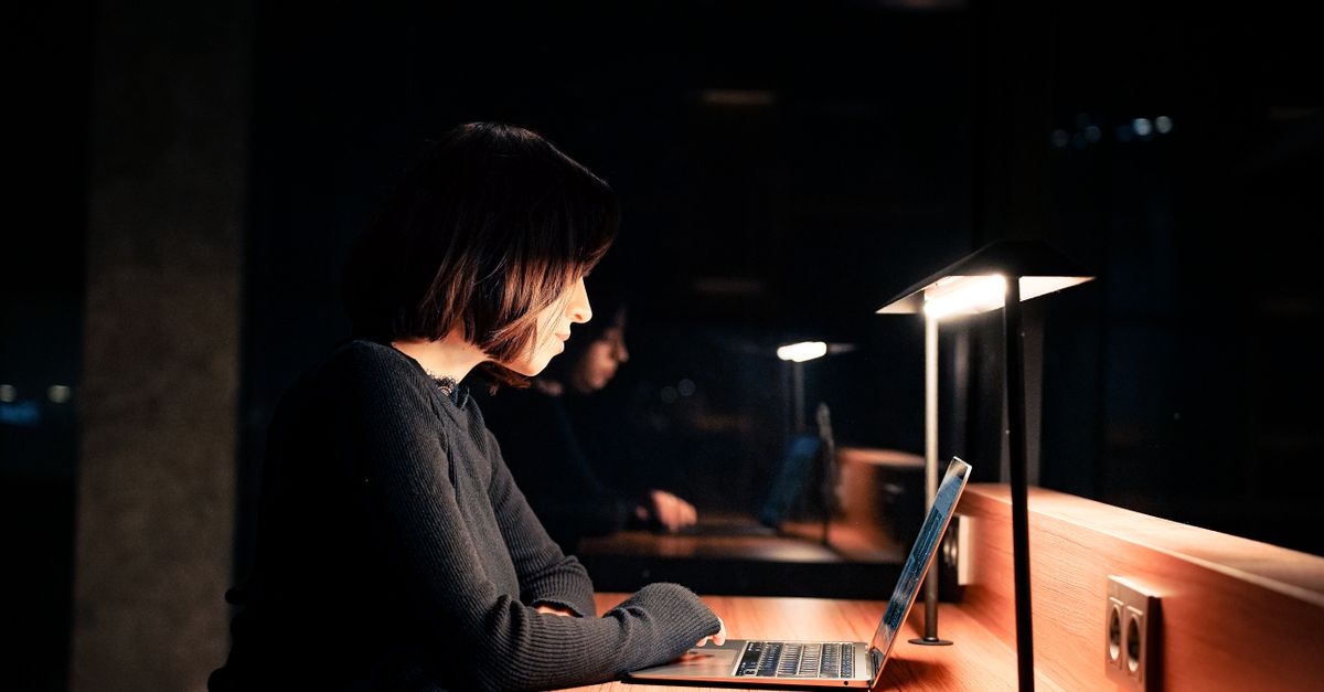 A woman focused on her laptop at a desk with dim lighting, working late at night.